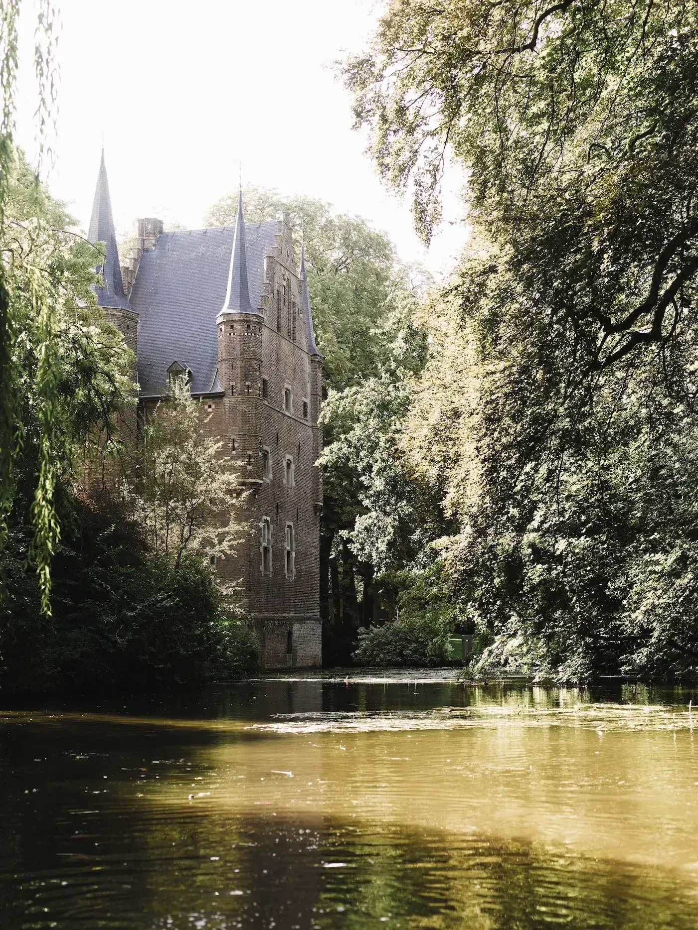 Kasteel Gemert outside with tall spires surrounded by lush trees, reflected in a calm, sunlit pond.
