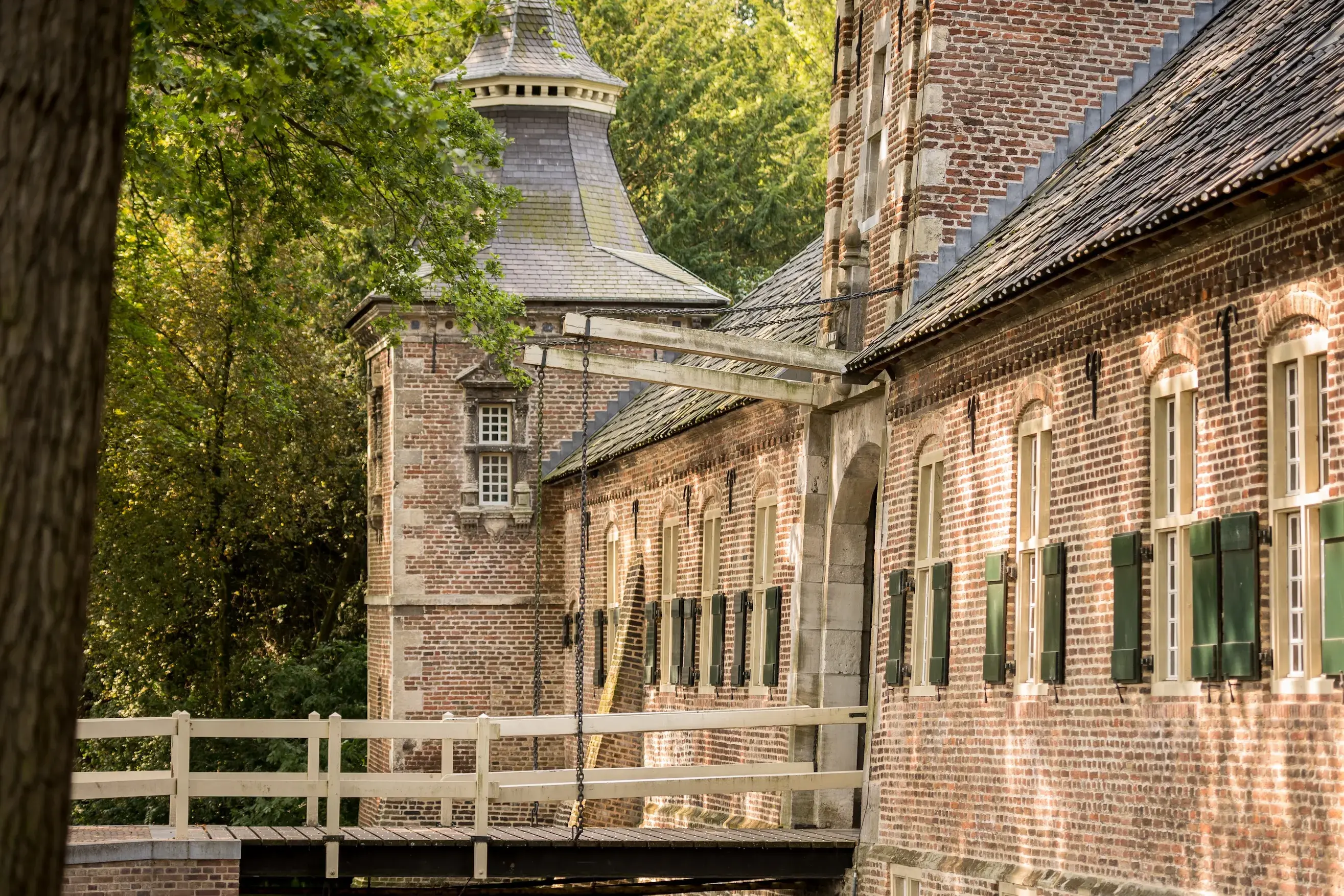 Historic brick building with green shutters and a wooden bridge, surrounded by trees, under a clear sky.