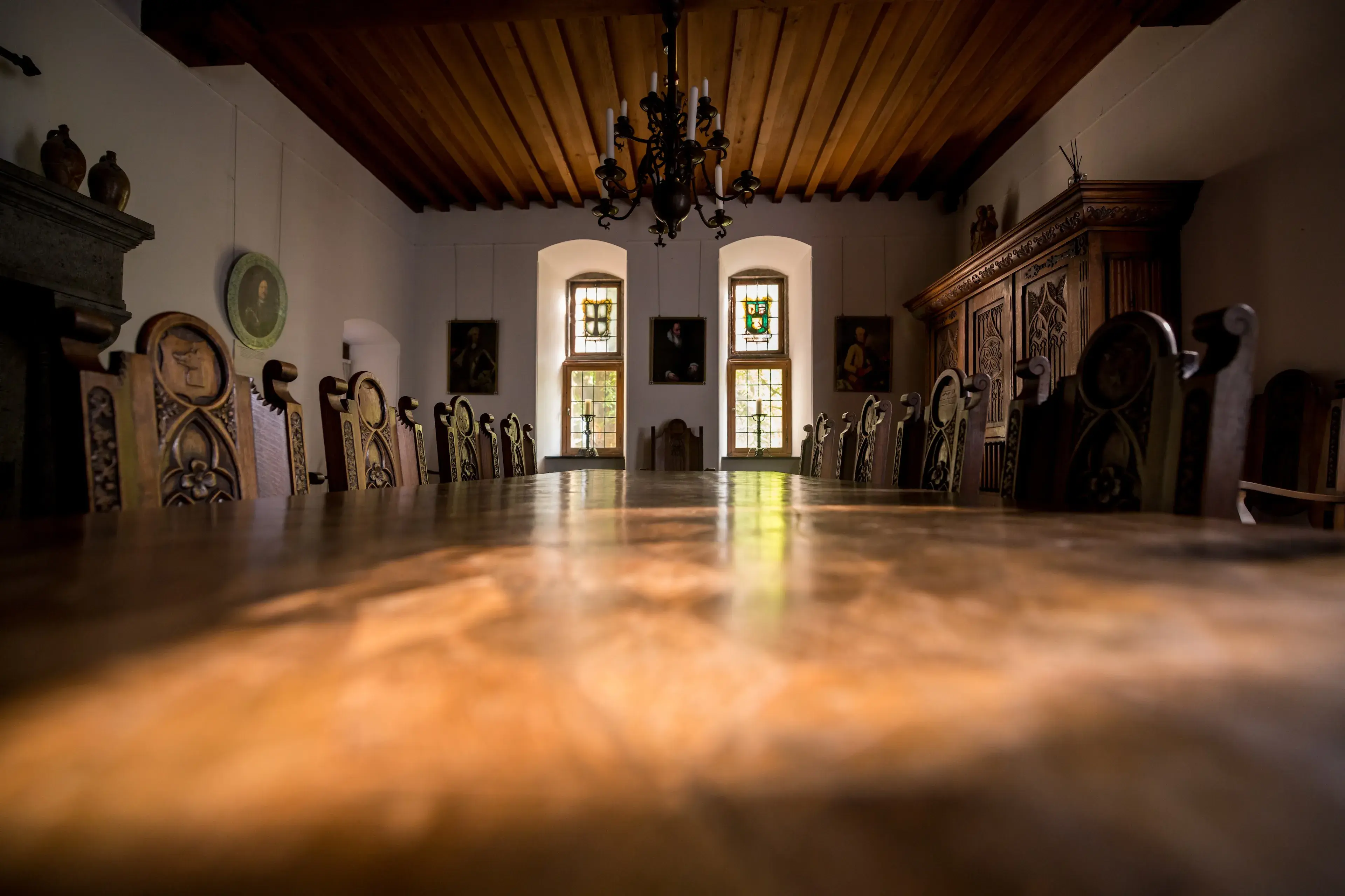 A dimly lit, rustic dining room with a long wooden table, ornate chairs, a chandelier, stained glass windows, and a wooden ceiling.